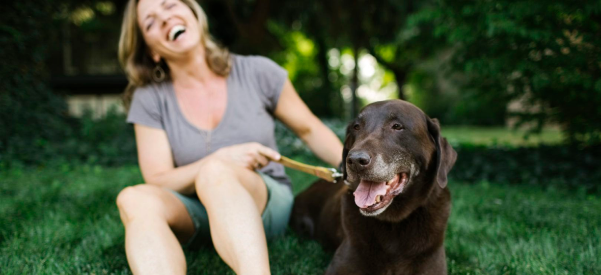 Laughing woman in sandals petting leashed chocolate Labrador retriever.